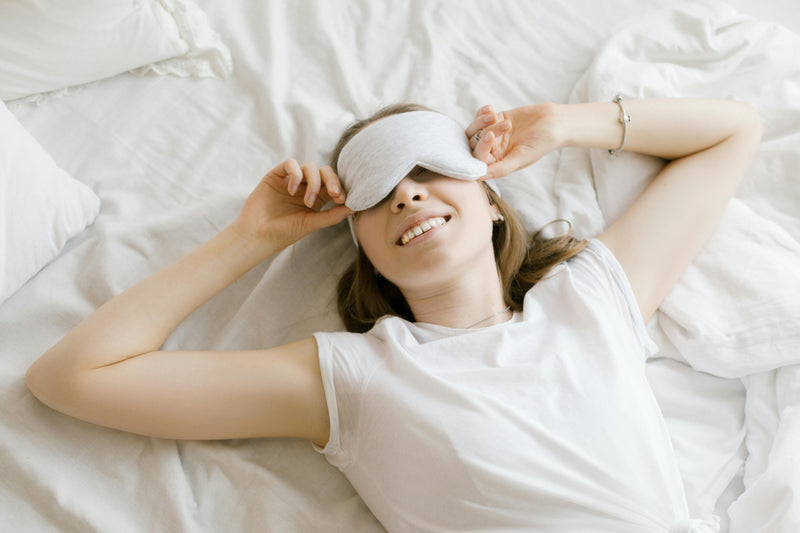 Mulher sorrindo com máscara de dormir em cama branca.
